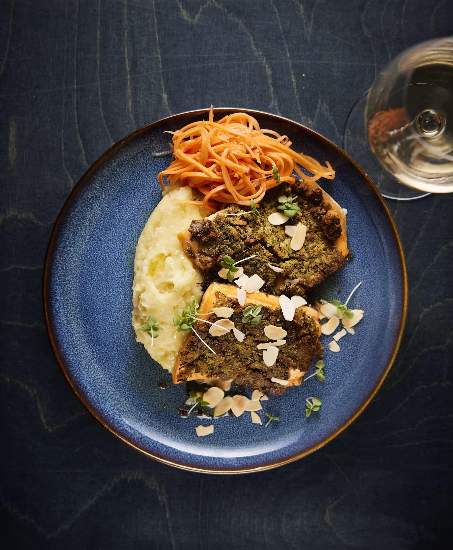 Herb-crusted fish with mashed potatoes, shredded carrots, microgreens, and sliced almonds on blue plate