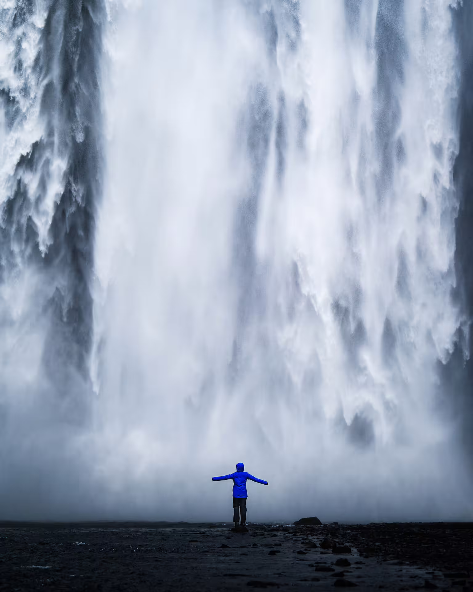 Person in blue jacket standing arms outstretched before massive Icelandic waterfall on guided Iceland tour