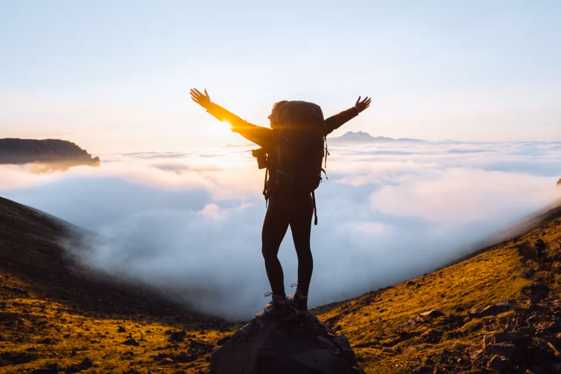 Hiker with backpack standing on rock above clouds at sunrise in Iceland mountains