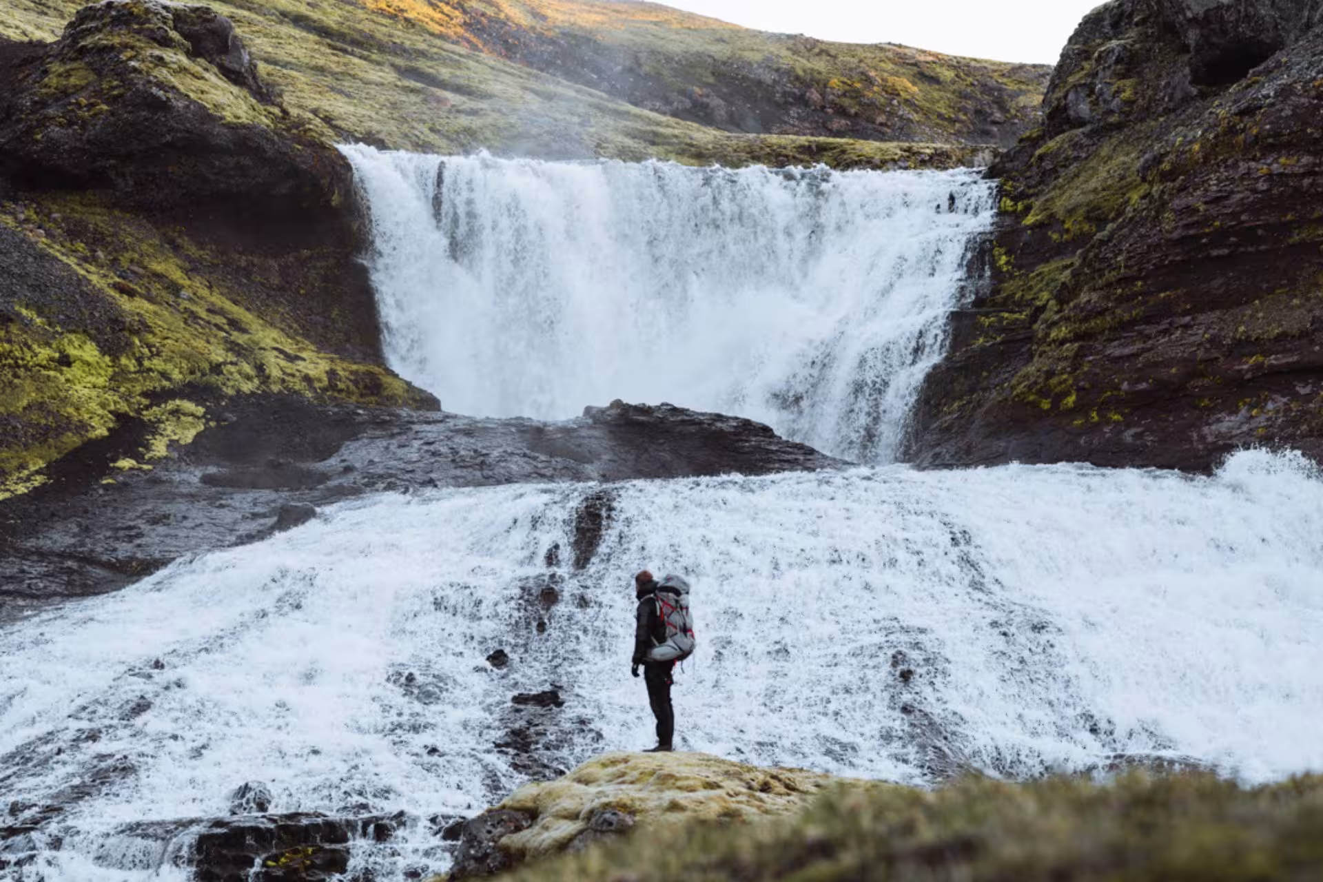 Hiker with backpack standing before powerful Icelandic waterfall surrounded by moss-covered lava rocks