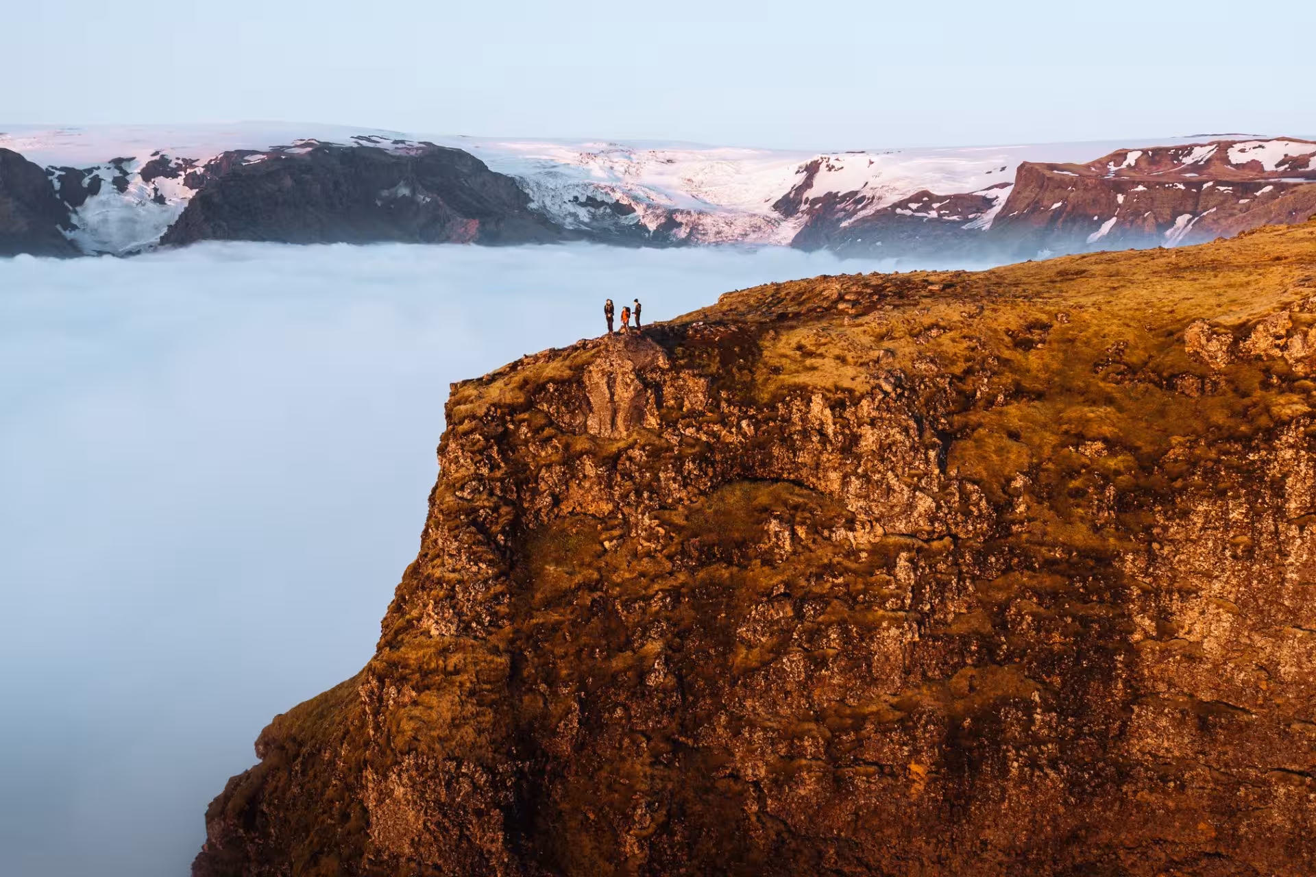 Three hikers standing on a dramatic cliff edge above clouds with snow-capped glaciers in Iceland