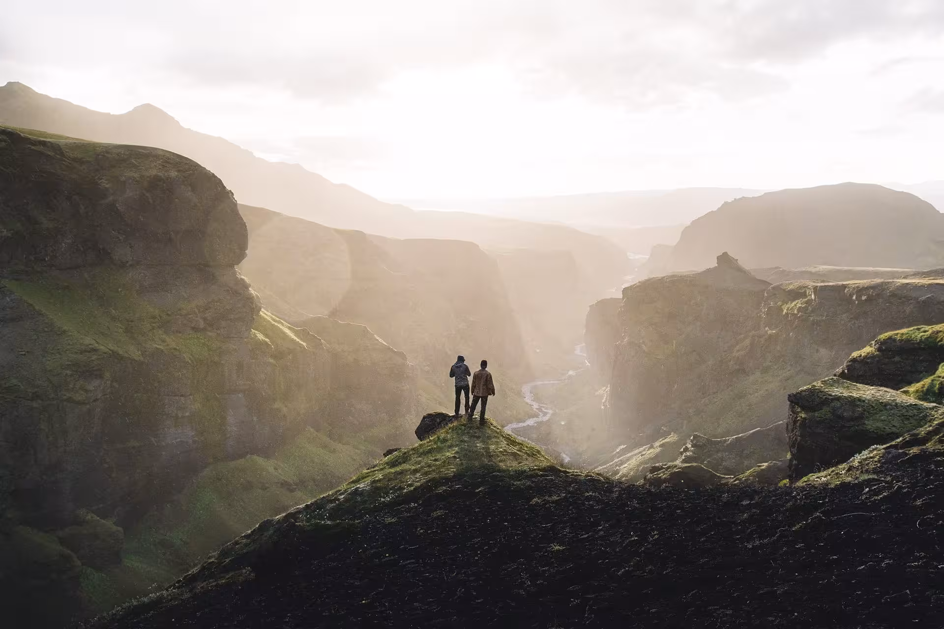 Two hikers standing on a mossy cliff overlooking a dramatic Iceland canyon valley with misty mountains