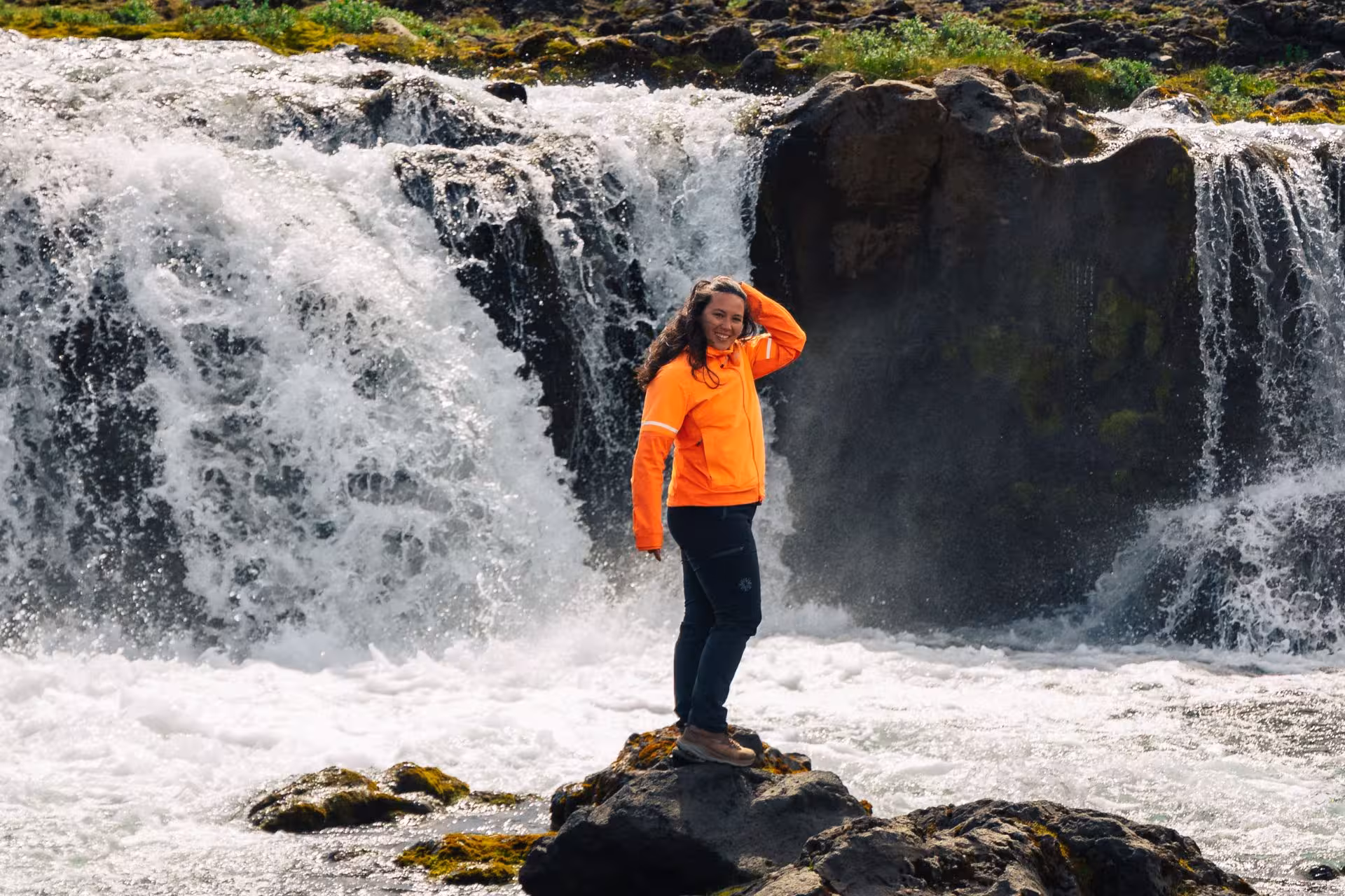 Smiling woman in orange jacket standing on rocks in front of powerful Iceland waterfall