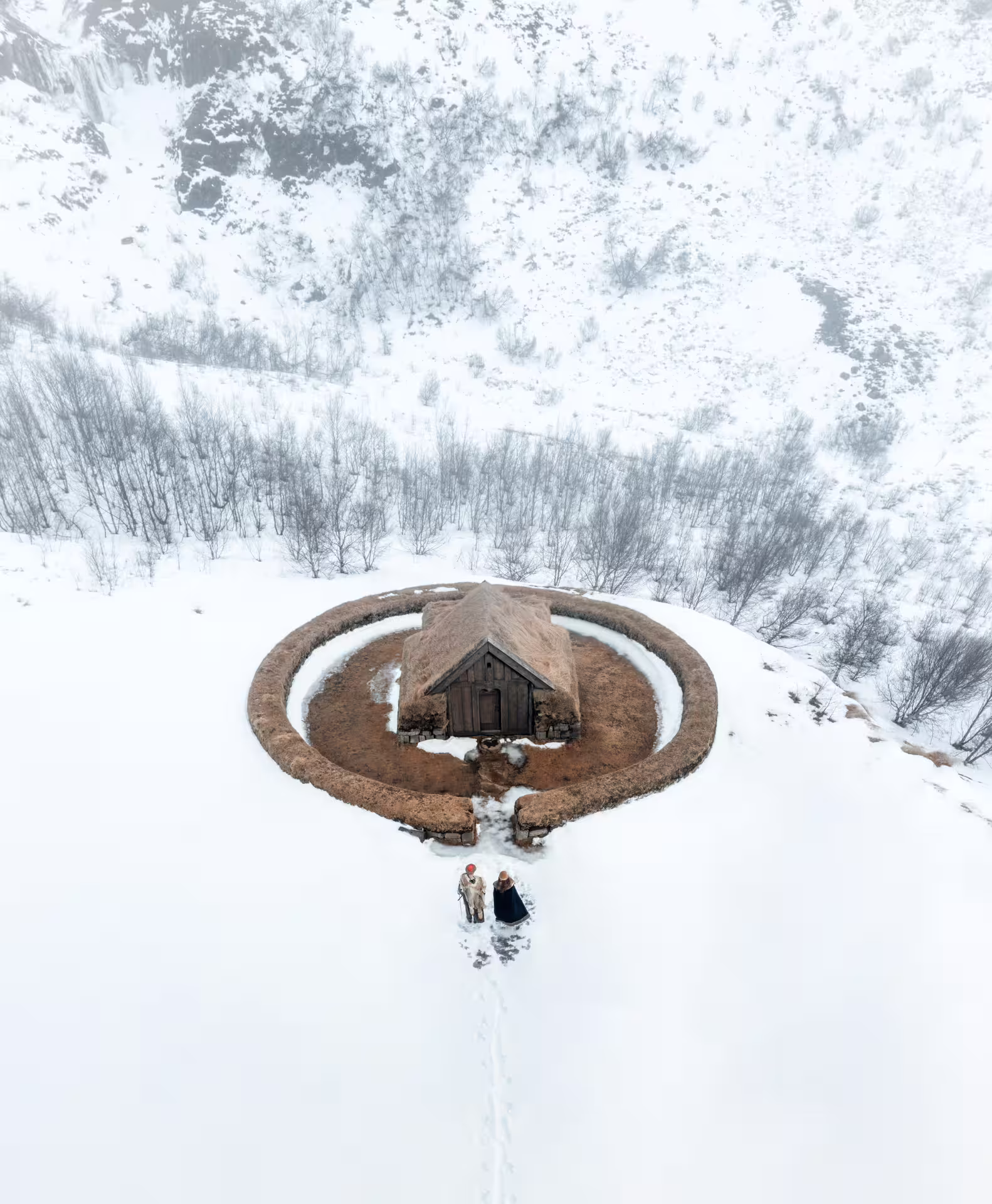 Aerial drone view of Iceland Viking circle longhouse surrounded by snow with two costumed Vikings approaching