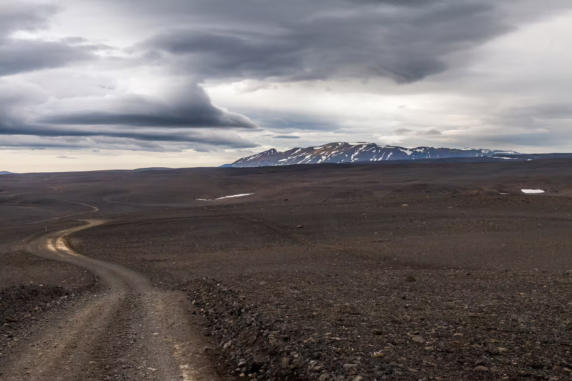Winding gravel road cutting through Iceland volcanic highland desert with snow-capped mountains on a 12-day grand tour.