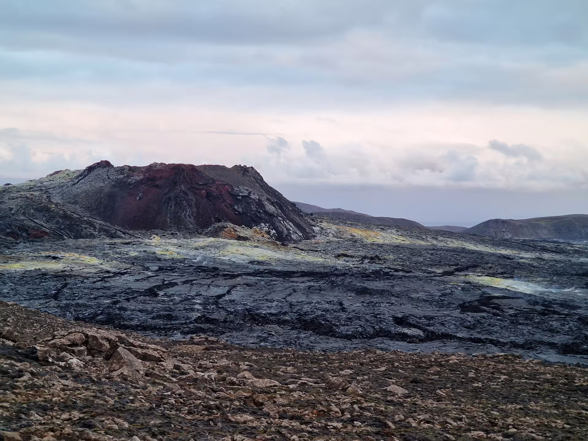 Solidified black lava field with red volcanic crater and sulfur deposits under cloudy sky in Iceland