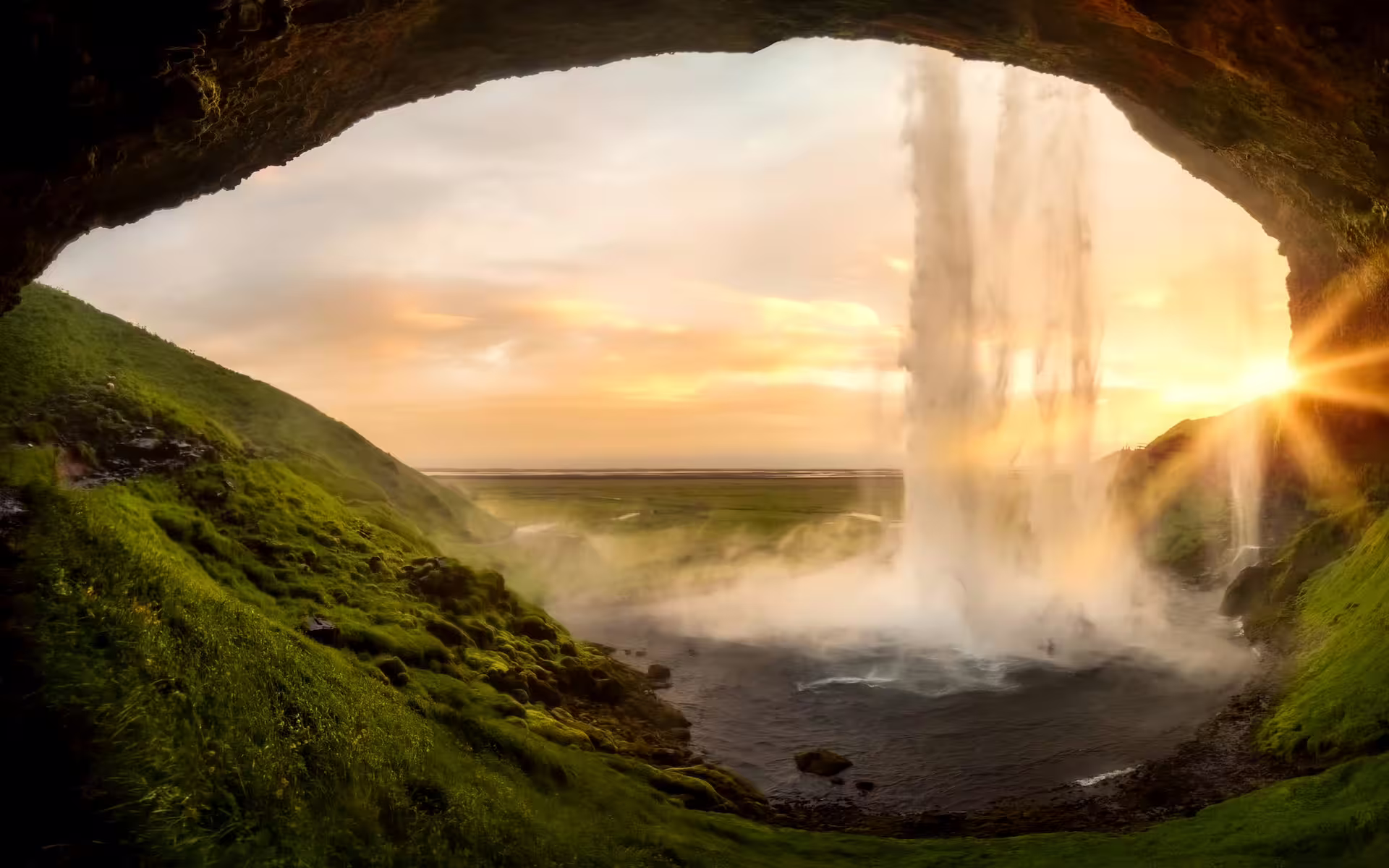 View from behind Seljalandsfoss waterfall in Iceland at golden sunset with misty spray and green hillside