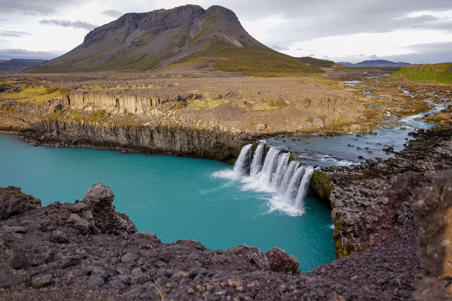 Stunning Iceland highlands waterfall cascades into vivid turquoise glacial river with dramatic volcanic mountain and basalt columns backdrop.