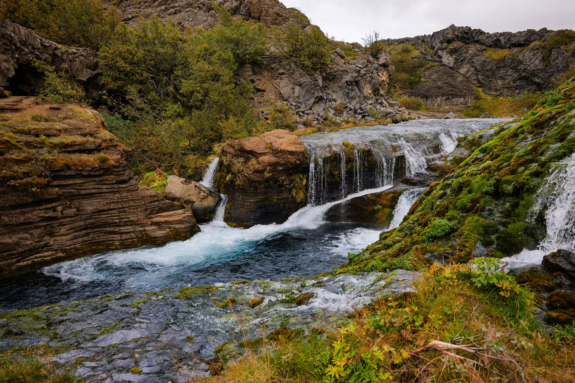 Crystal-clear cascading waterfall rushing over layered rocks through a lush mossy Iceland highland canyon gorge.
