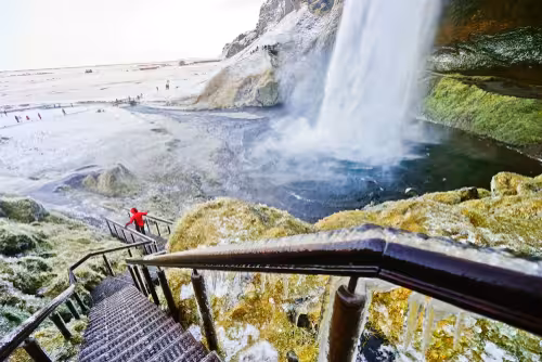 Visitor in red jacket on icy stairs beside Seljalandsfoss waterfall in Iceland winter