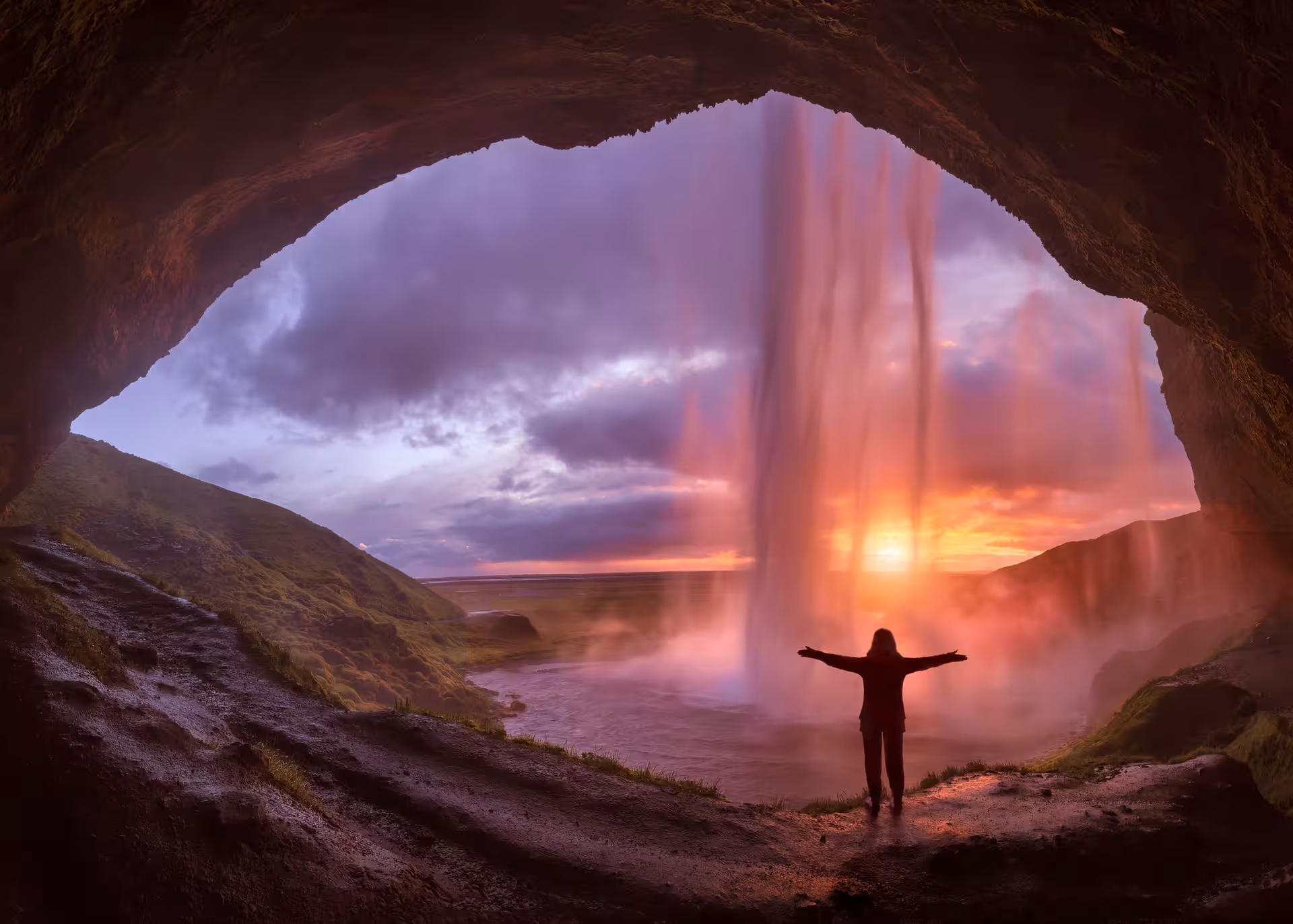 Person standing behind Seljalandsfoss waterfall at sunset in Iceland, arms outstretched inside cave