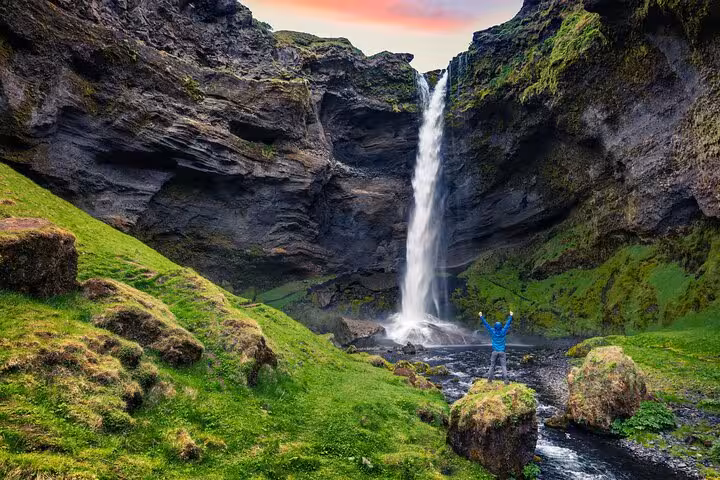 Person standing arms raised before tall Iceland waterfall cascading through mossy green canyon cliffs