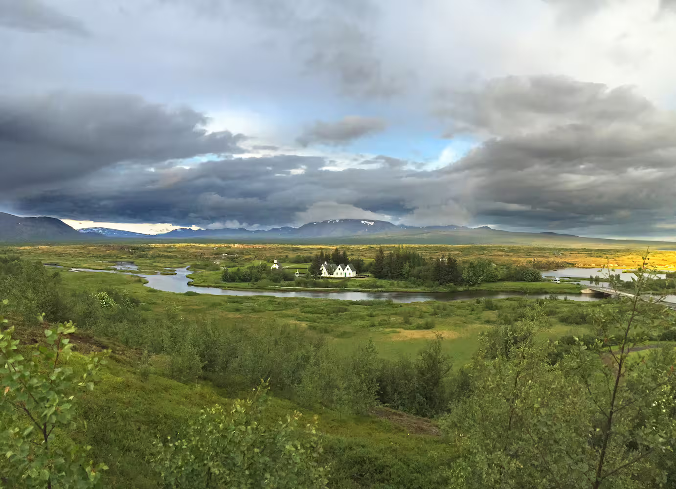 Panoramic view of Iceland's green valley with winding river, white farmhouses, and snow-capped mountains under dramatic sky