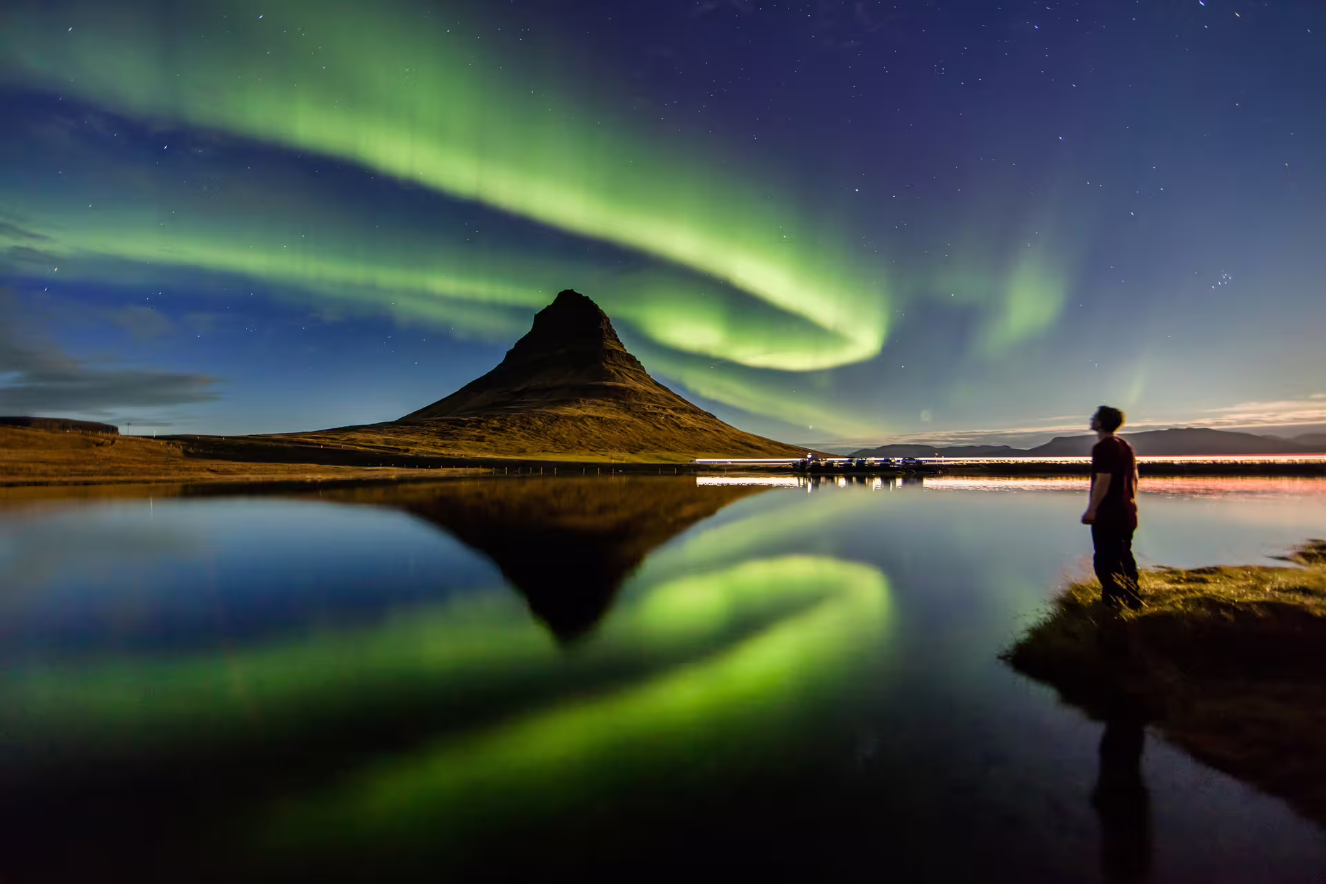 Person watching aurora borealis over Kirkjufell mountain reflected in calm lake, Iceland winter night
