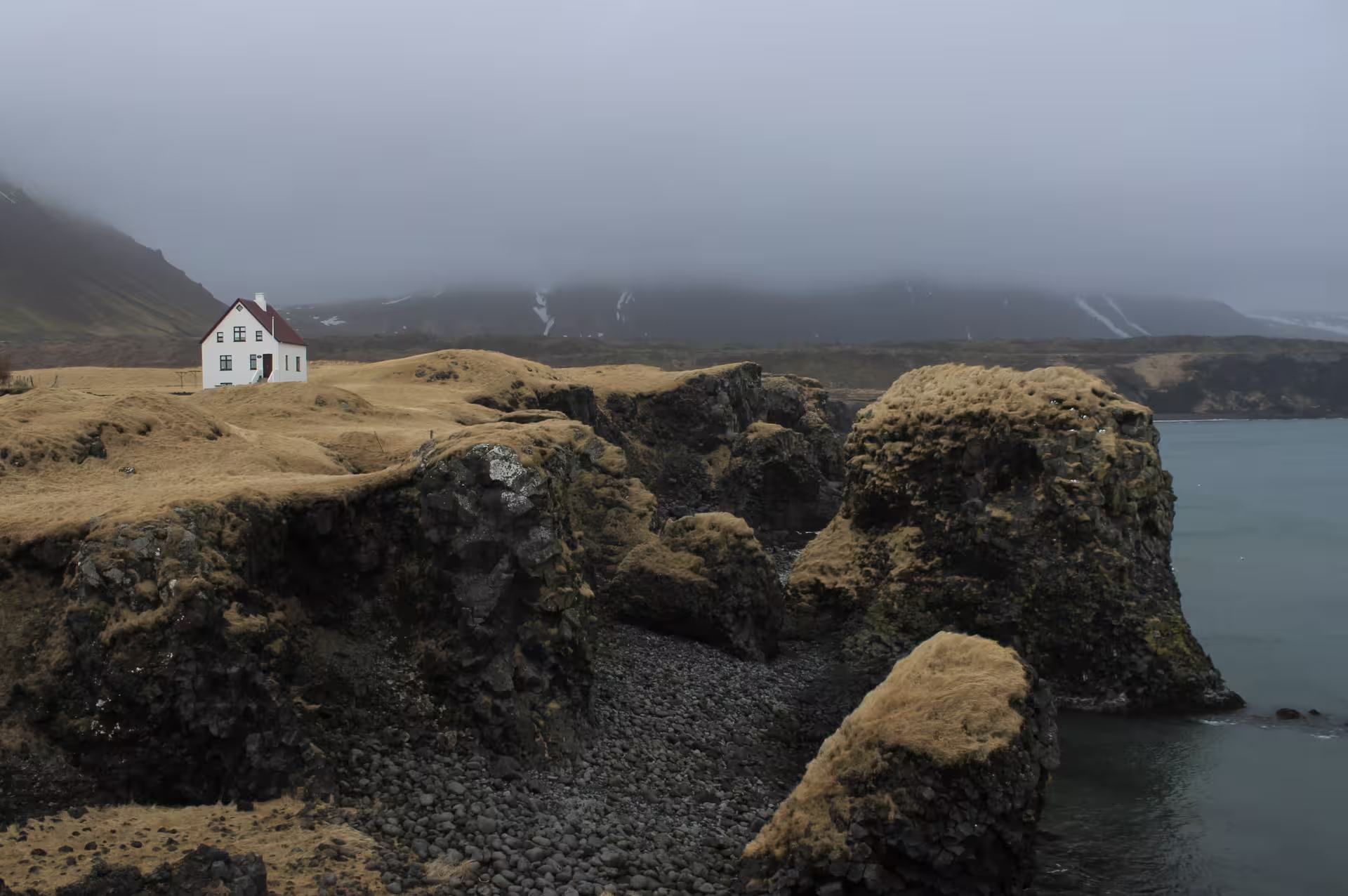 Lone white house with red roof on mossy volcanic cliffs above Iceland's rocky coastline in foggy winter weather