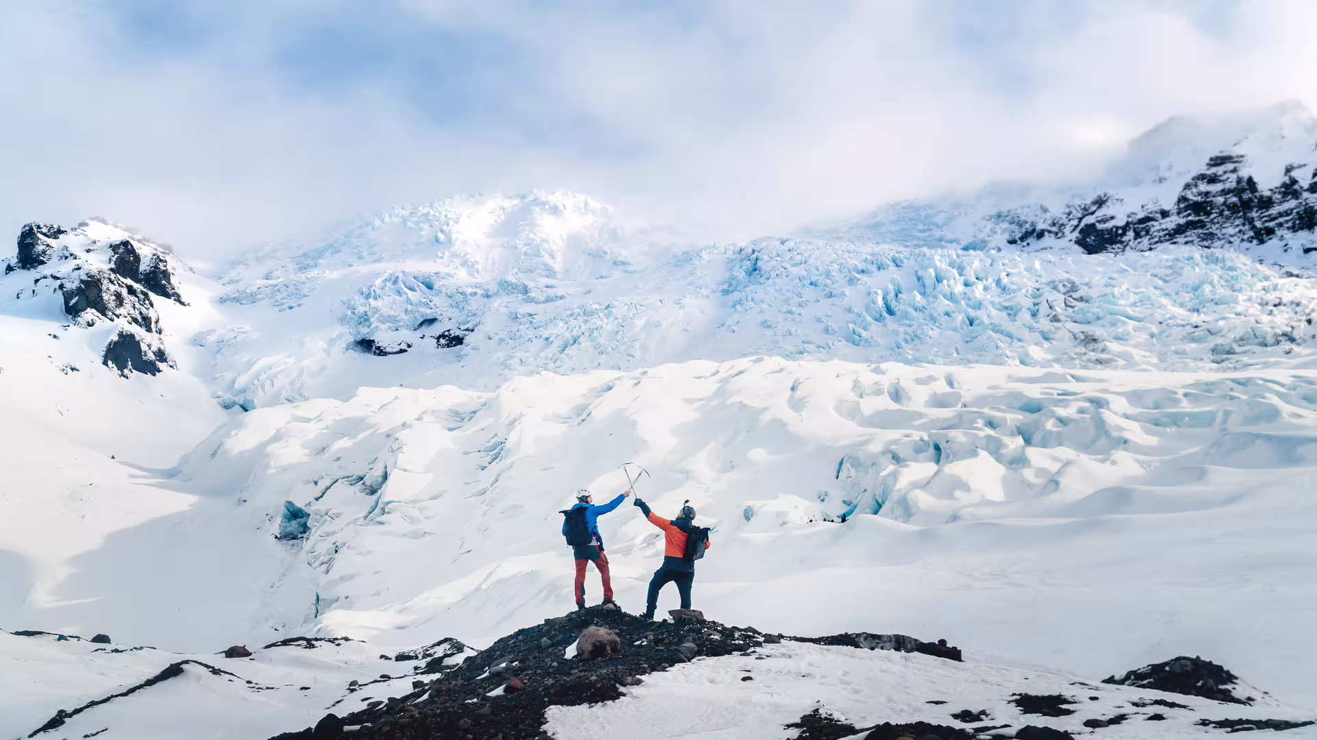 Two climbers celebrating with ice axes on a rocky outcrop in front of a vast Icelandic glacier