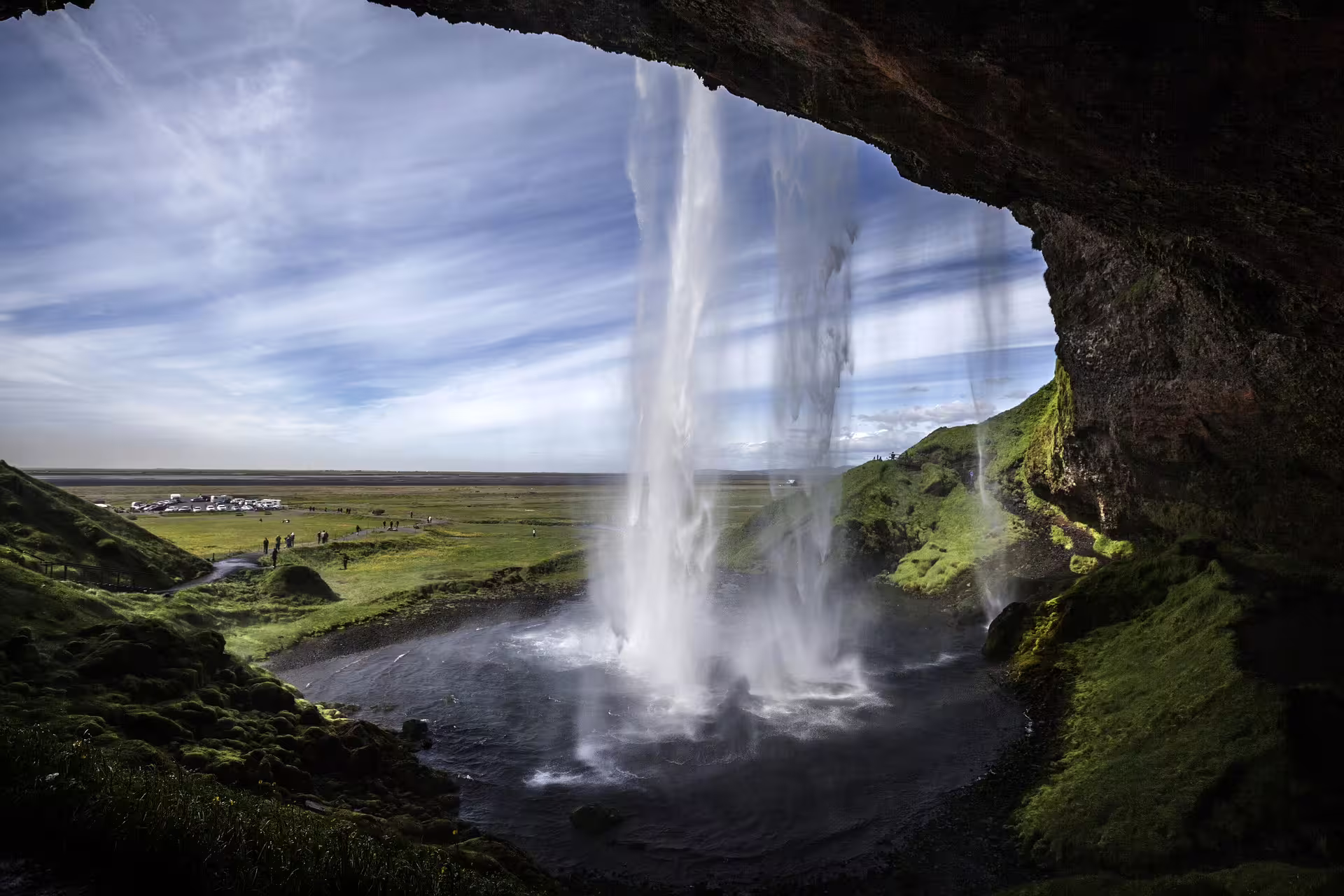 View from behind Seljalandsfoss waterfall in Iceland, with green moss-covered cliffs and vast plains beyond