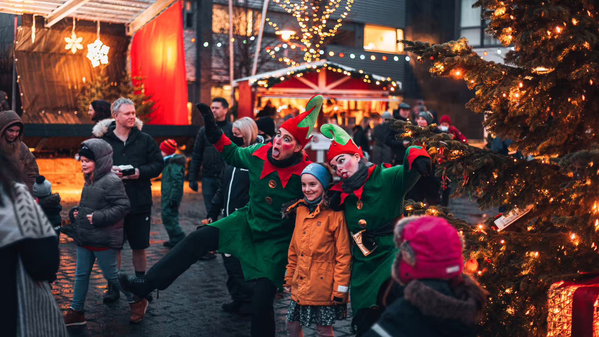 Two elves in green costumes posing with a child at an Icelandic Christmas market with festive lights