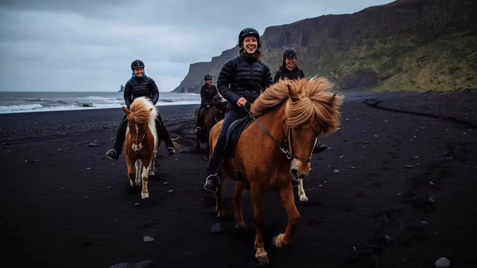 Group of riders on Icelandic horses along a black sand beach with dramatic cliffs in background