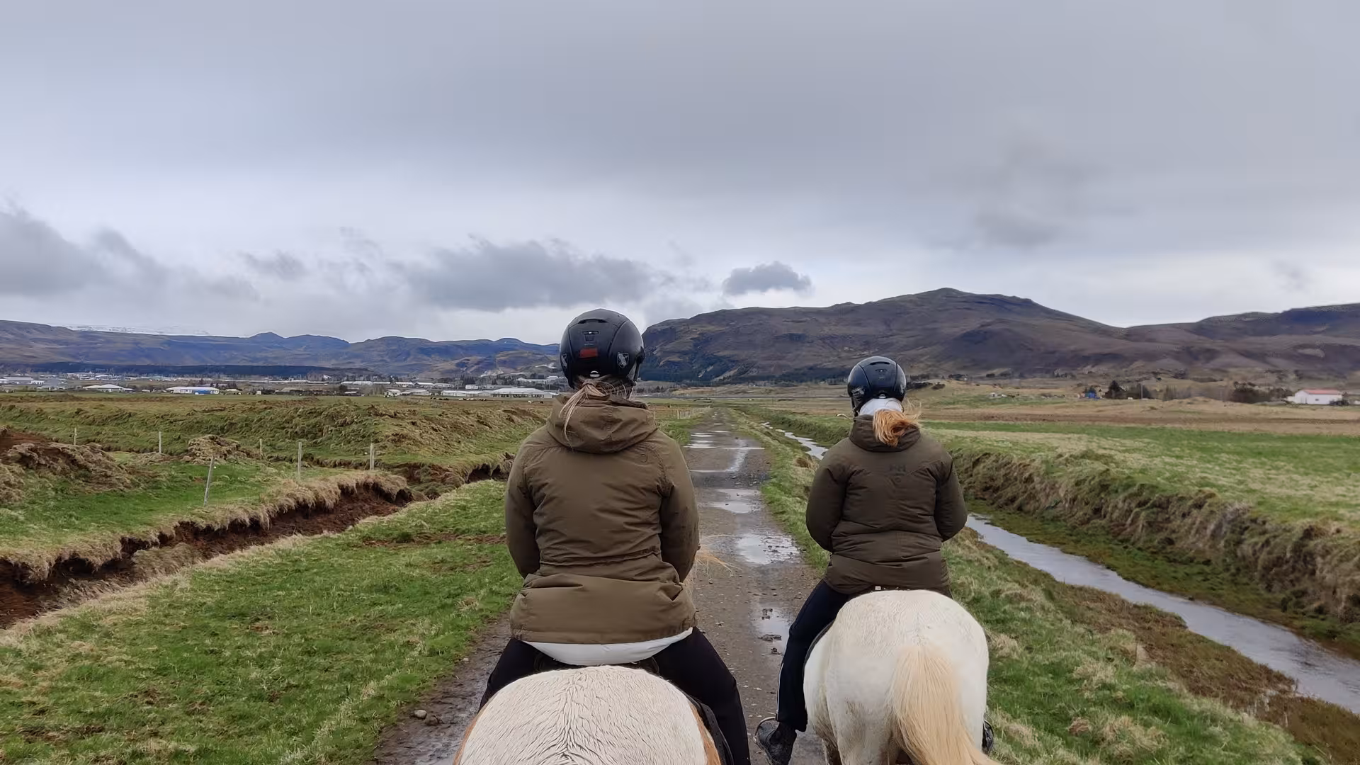Two riders on white Icelandic horses trekking a scenic countryside trail with misty mountains backdrop