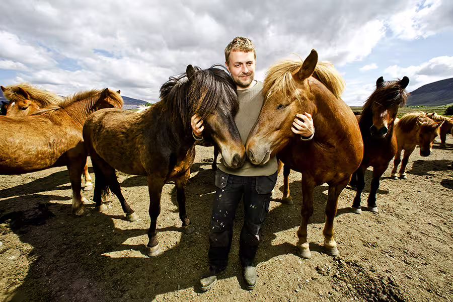 Man embraces two Icelandic horses under a cloudy sky, showcasing the horseback riding experience on the Silfra Snorkeling tour.