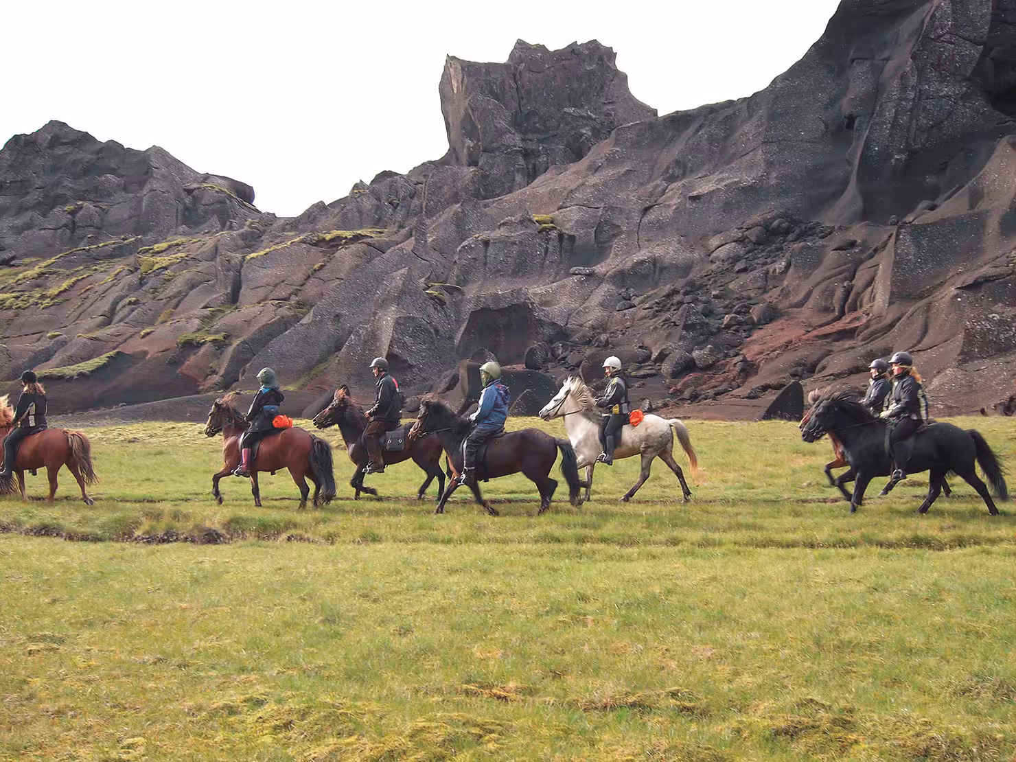 Riders on Icelandic horses crossing green meadow beneath volcanic cliffs on Marardalur Valley of Horses tour