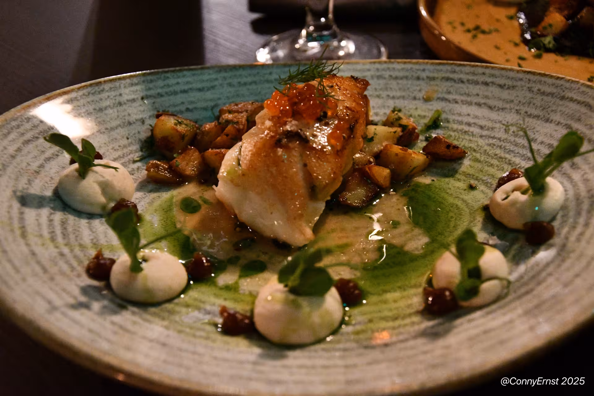 Pan-seared white fish with salmon roe, roasted potatoes, green herb sauce and cream dollops on ceramic plate