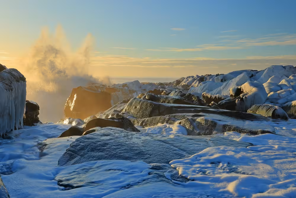 Snow-covered rocky shoreline with crashing waves and golden sunrise light in Iceland's winter landscape
