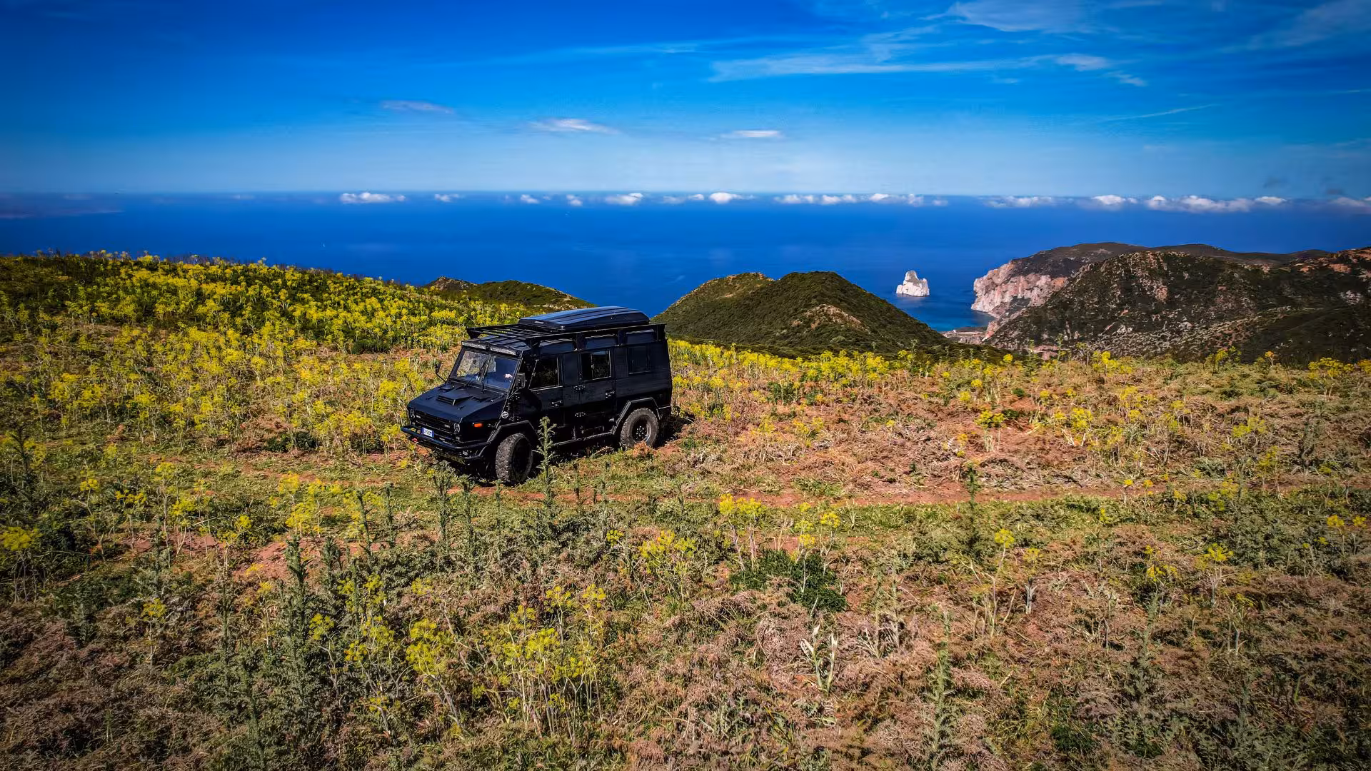 A rugged jeep traverses a scenic hilltop trail with panoramic views of Iglesias' coastline and lush greenery.