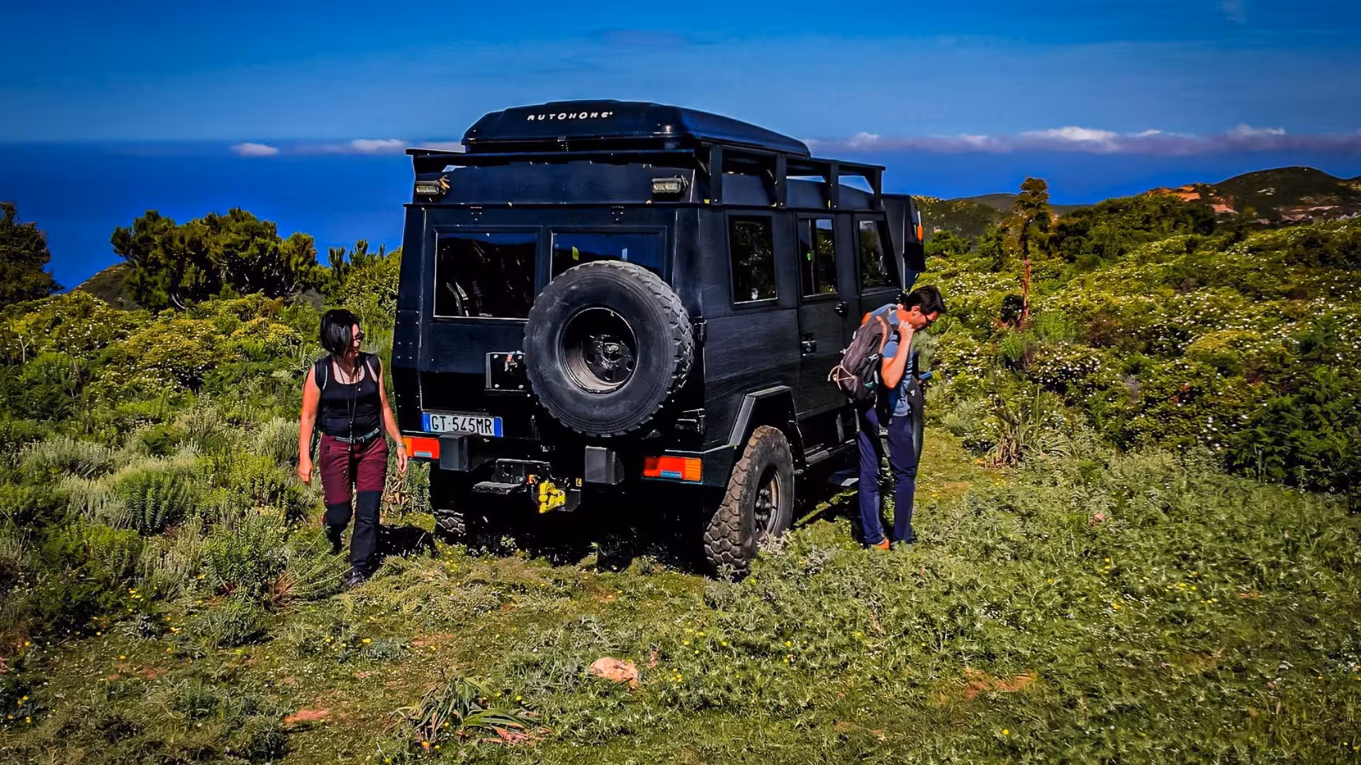 Travelers preparing for trekking near jeep on a scenic Iglesias tour, with ocean backdrop and green landscape.