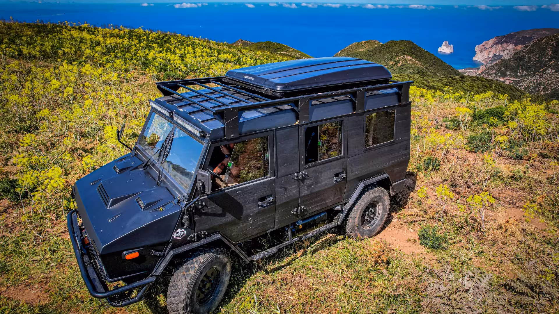 Close-up of a black jeep navigating through vibrant wildflower fields with stunning sea views in Iglesias.