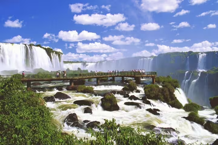 Scenic view of Iguazu Falls with tourists on a walkway, showcasing the majestic waterfalls under a vibrant blue sky.