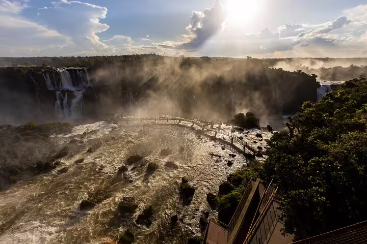 Breathtaking view of Iguazu Falls at sunset with mist rising and a scenic walkway, perfect for a guided adventure tour.