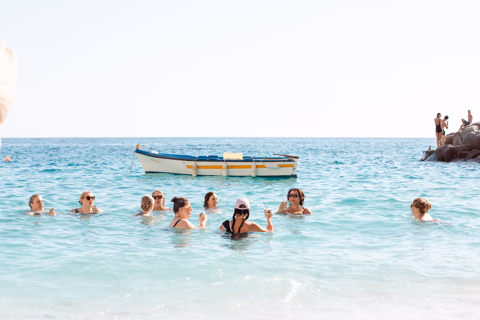 Group of friends enjoying Ikaria’s Therma hot springs in the sea, chatting with drinks beside a traditional Greek fishing boat