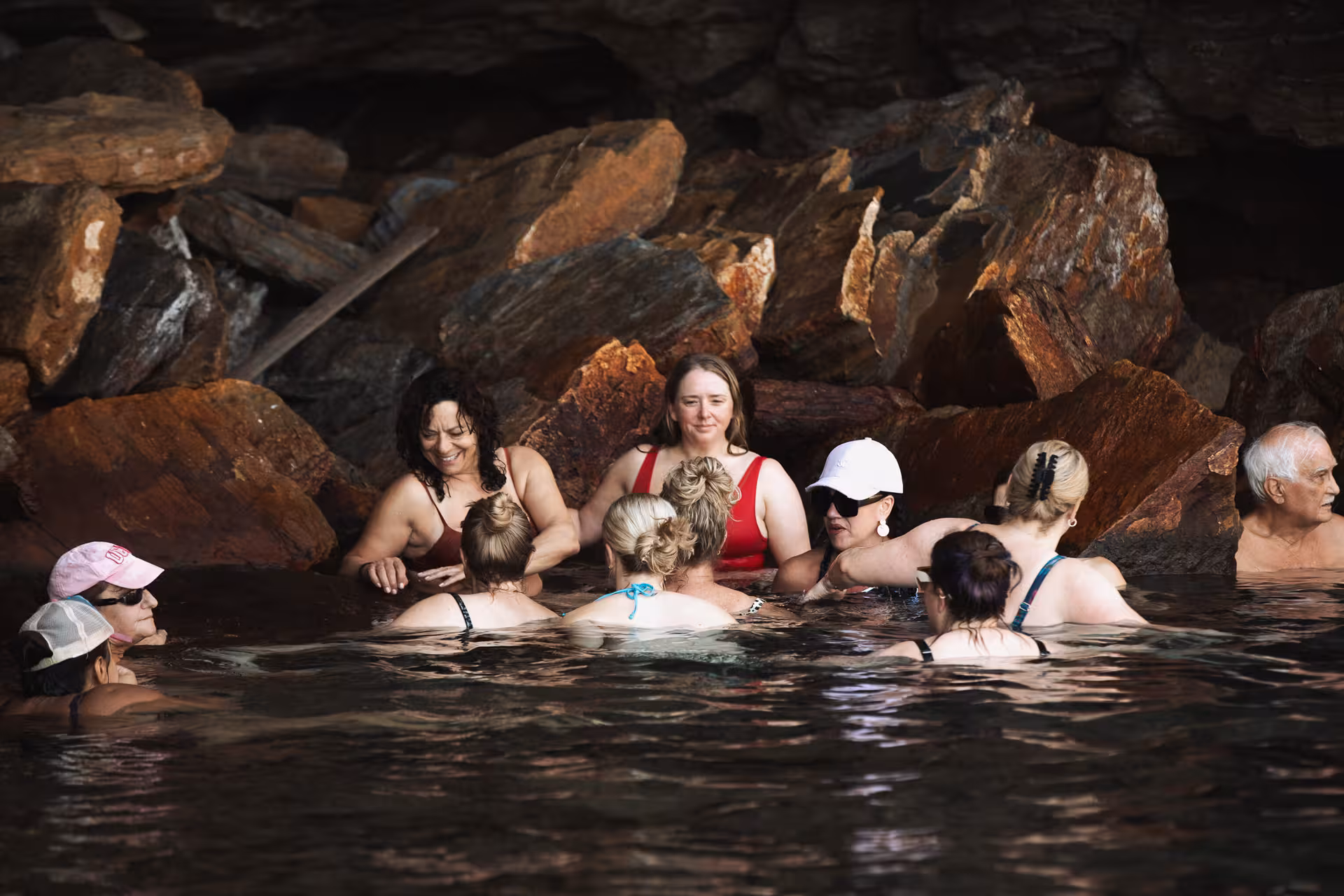 Group of visitors soaking in Ikaria’s therapeutic thermal springs by rugged rocks, a highlight of the island’s wellness tours