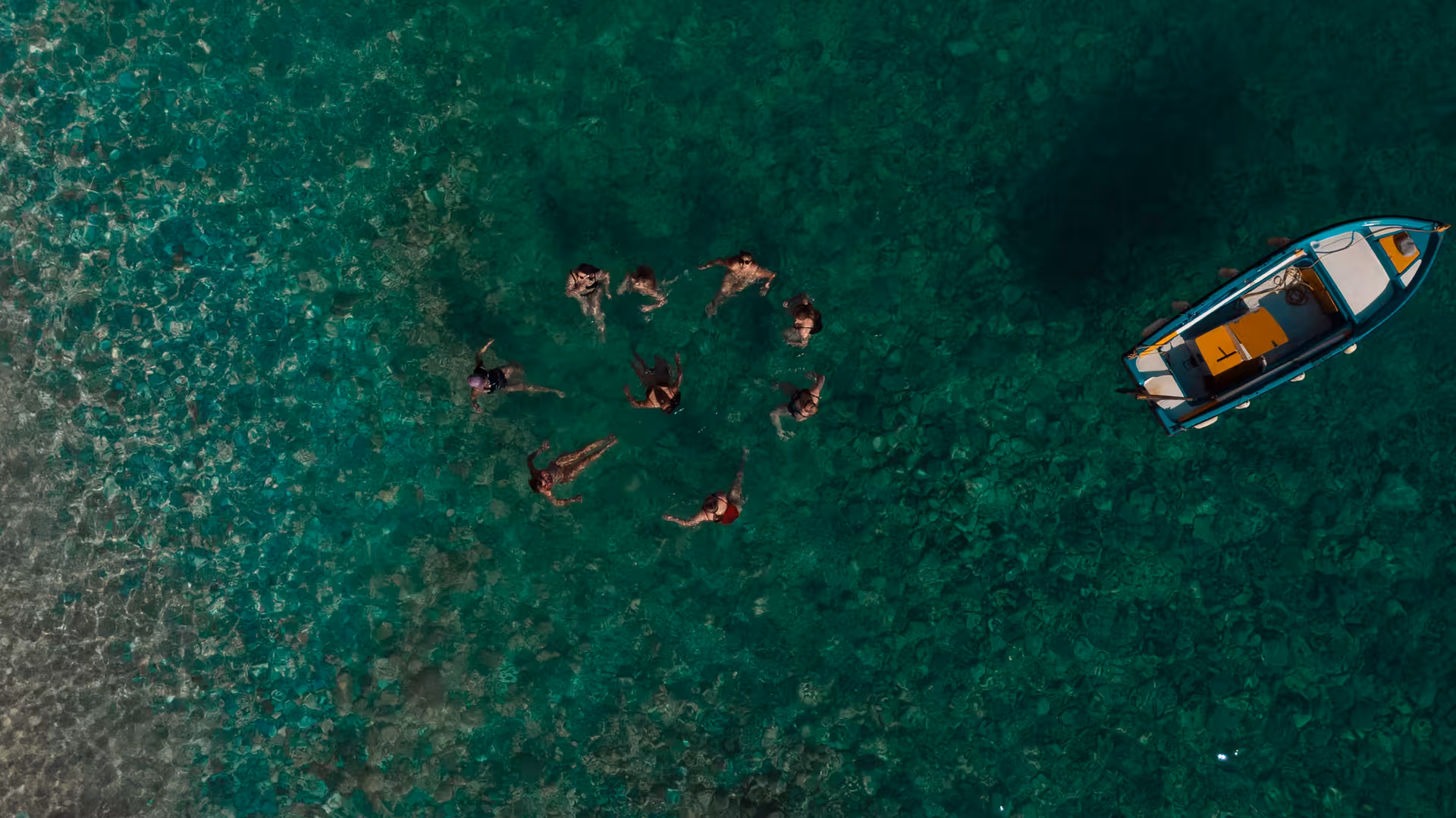 Group of friends swimming in clear turquoise Ikaria waters near a small boat during Aegean coastal excursion