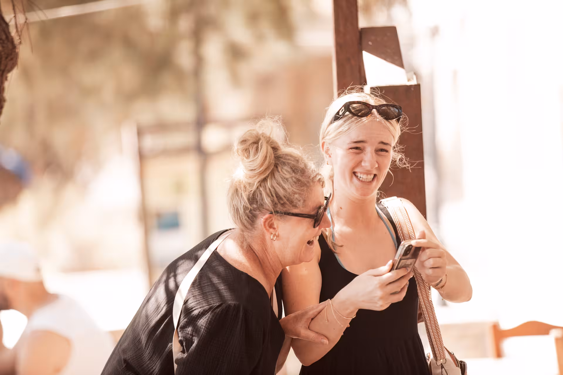 Two women laugh together while checking a phone during an Ikaria village stop, capturing joyful moments on the island tour