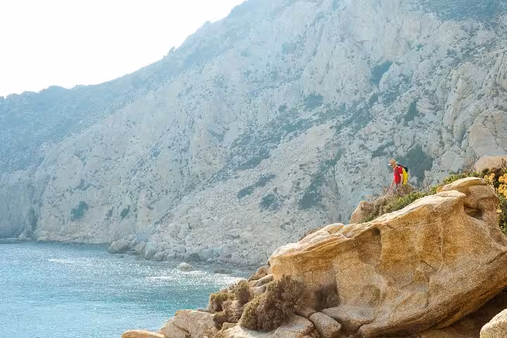 Hiker stands on sunlit cliffs above the turquoise Aegean Sea on Ikaria’s wild west coast, exploring secluded beaches.