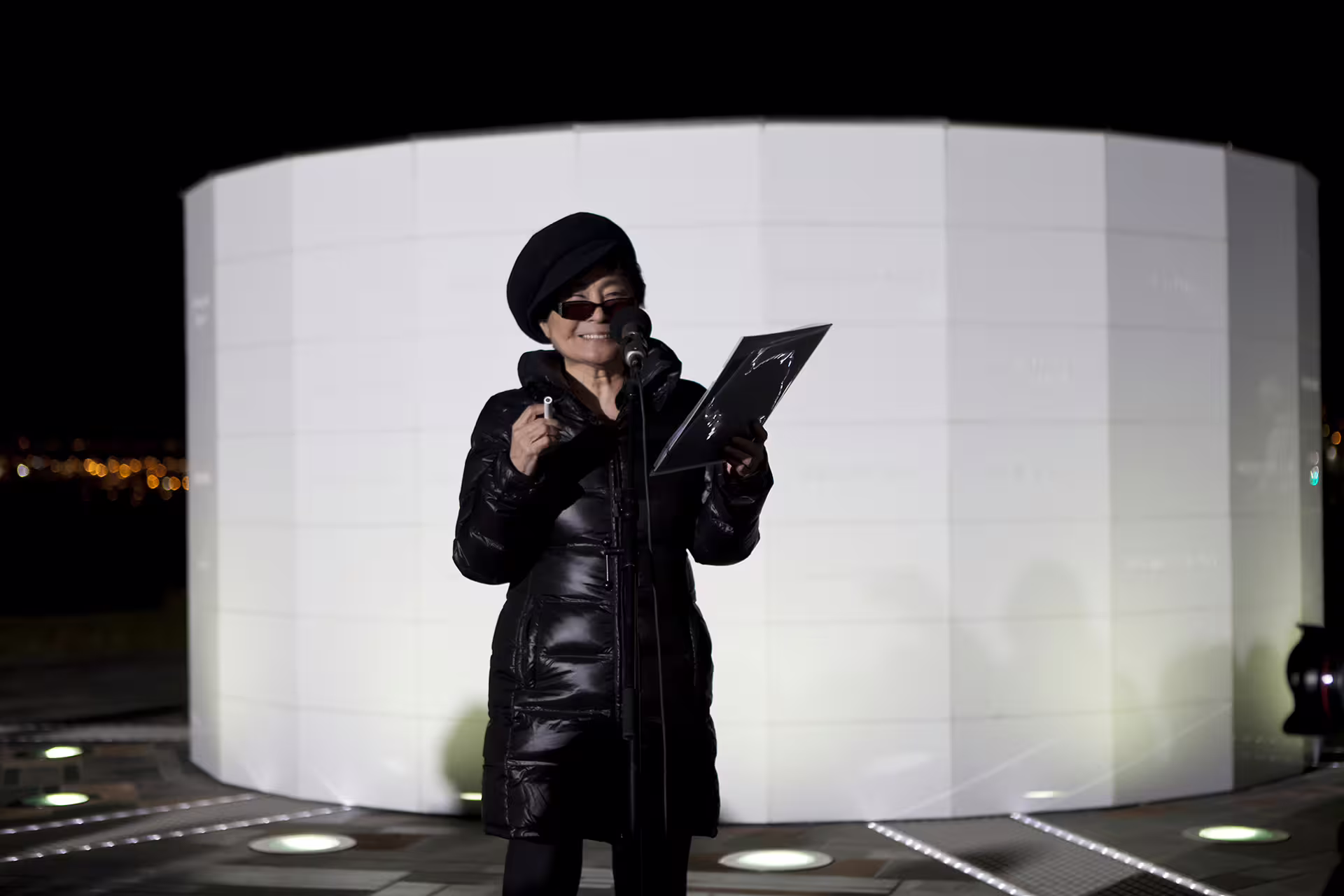 Woman in black coat and beret speaking at microphone beside illuminated Imagine Peace Tower at night
