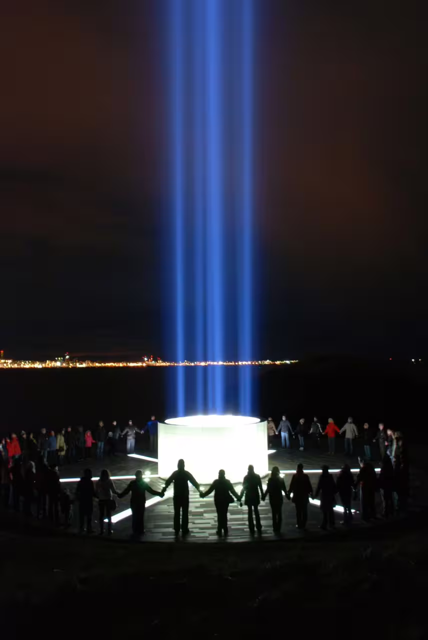 People holding hands in a circle around the illuminated Imagine Peace Tower sending blue light beams into the night sky on Videy Island