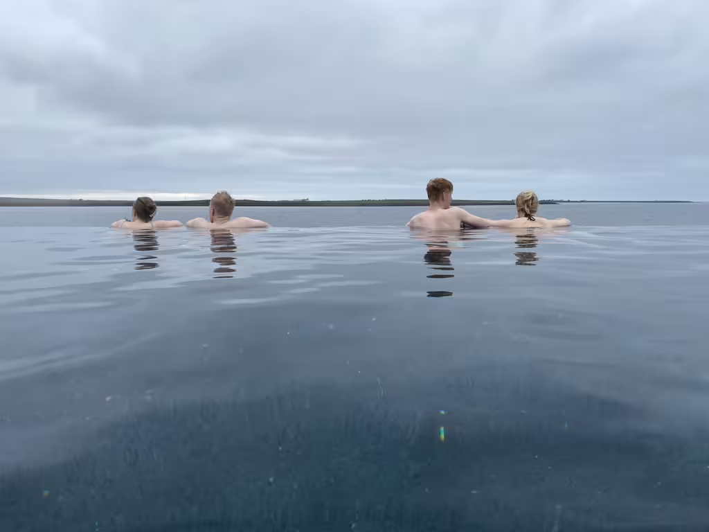 Four guests relaxing in an infinity geothermal pool overlooking Iceland's serene landscape under cloudy skies