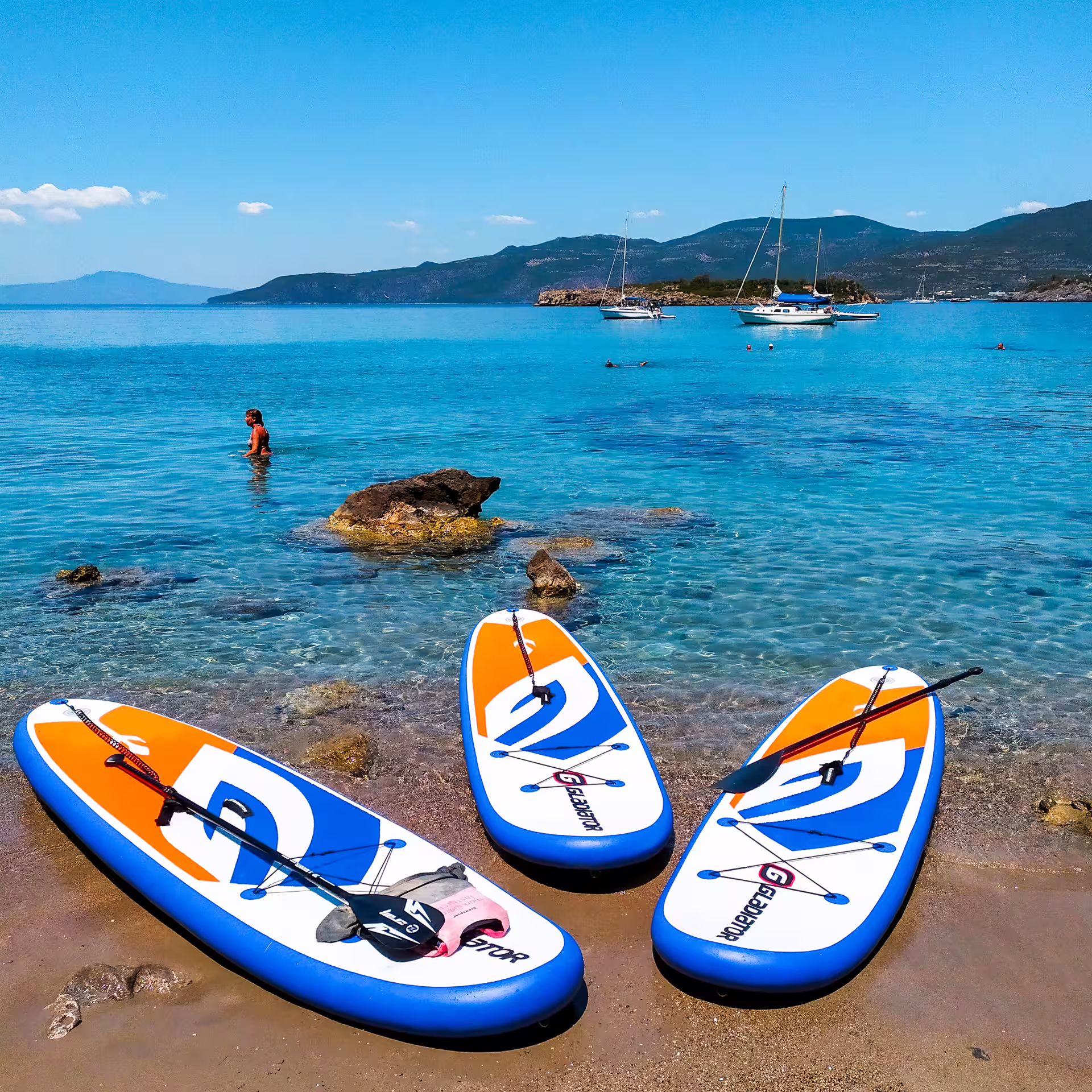 Inflatable SUP boards ready on a sandy beach in Kalamata with calm blue bay, rocks and sailboats
