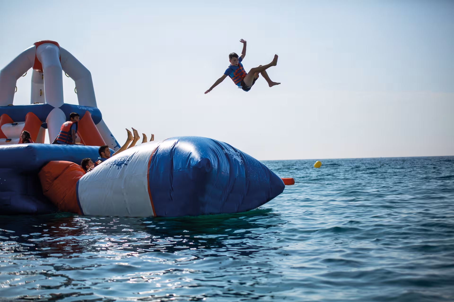 A kid launches high into the air from an inflatable waterpark blob launcher over the open ocean on a sunny day.