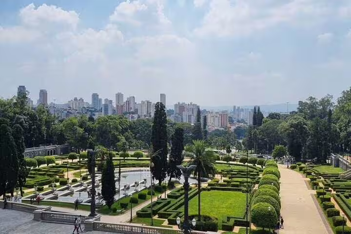 View of Ipiranga Museum gardens in São Paulo, showcasing manicured hedges and city skyline in the background.