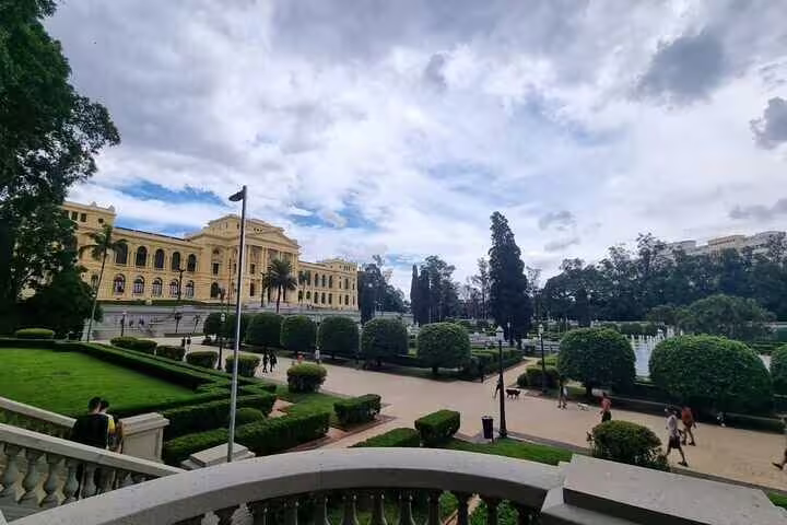 Scenic view of the Ipiranga Museum's neoclassical architecture and lush gardens under a partly cloudy sky.
