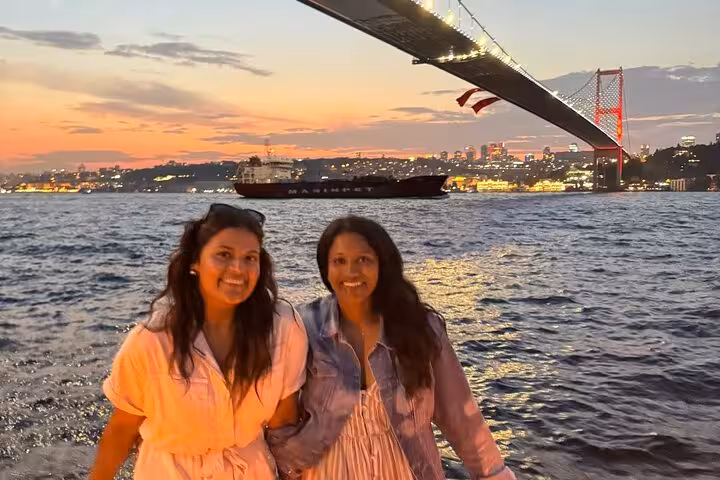 Two women posing on a Bosphorus luxury yacht at sunset with Istanbul lights and the Bosphorus Bridge behind