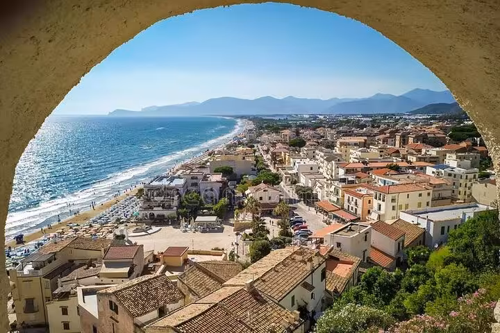 Panoramic coastal Italian town with terracotta rooftops, sandy beach, turquoise sea, and mountain backdrop viewed through stone arch