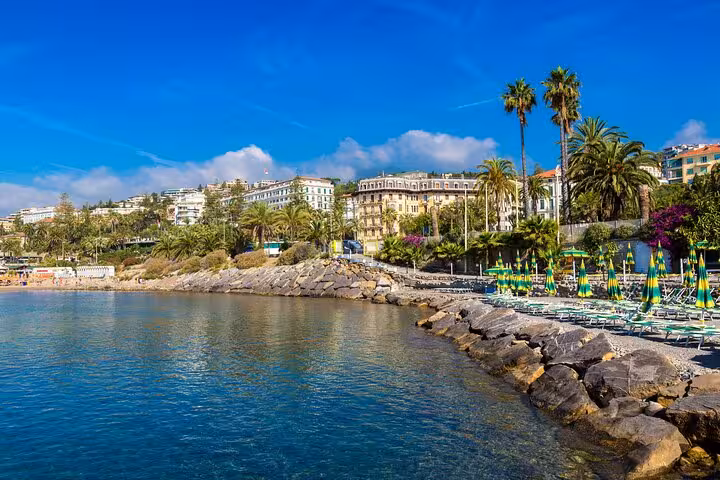 Scenic view of the Italian Riviera coastline with palm trees and vibrant beach umbrellas against a clear blue sky.