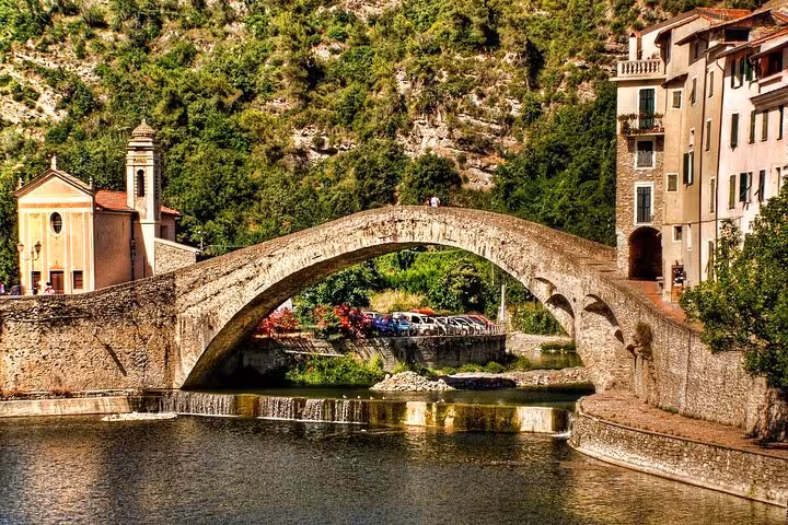 Ancient stone arch bridge over calm river in Italian Riviera village with church and hillside buildings