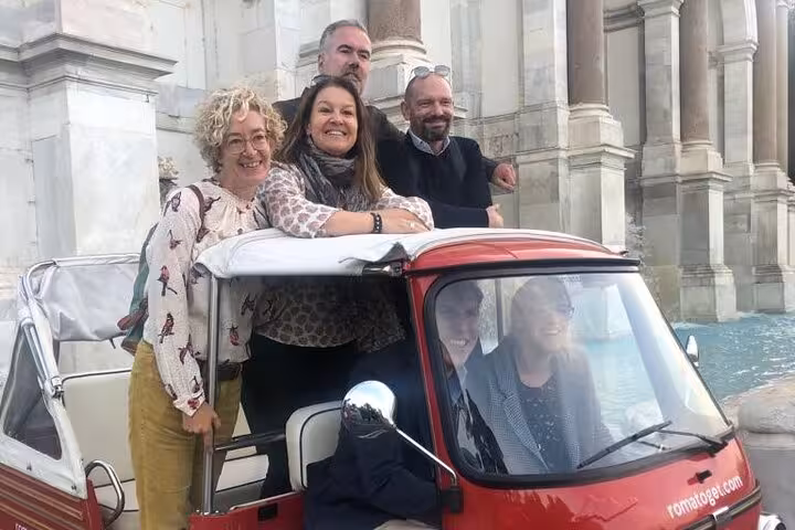 Group of friends pose on an Ape Calessino during a James Bond-themed tour, showcasing Rome's iconic architecture.